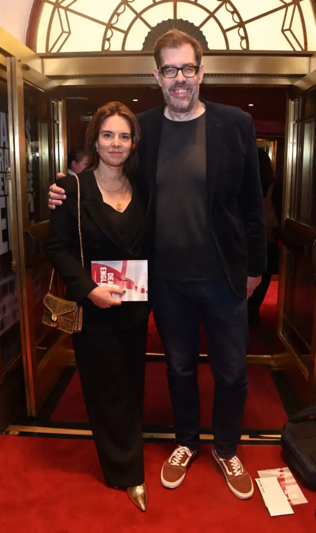 Ingrid and Richard stand on a red carpet; she wears a long dress and holds a booklet, he is in a casual outfit with glasses and sneakers. She comes up to his chin