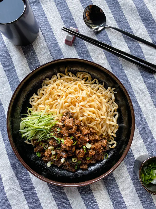 A bowl of noodles with diced meat, cucumber strips, and chopped green onions, set on a striped tablecloth with chopsticks nearby
