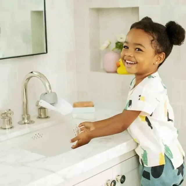Child smiling while washing hands with a faucet extender in a bathroom setting, showcasing a practical accessory for kids