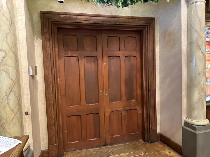 A pair of closed wooden double doors with decorative panels, framed by marble columns and topped with greenery.