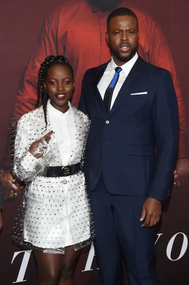 Lupita and Winston pose together; she wears a studded sheer jacket over a light outfit, and he wears a suit with a shirt and tie. Lupita is slightly taller than his shoulder