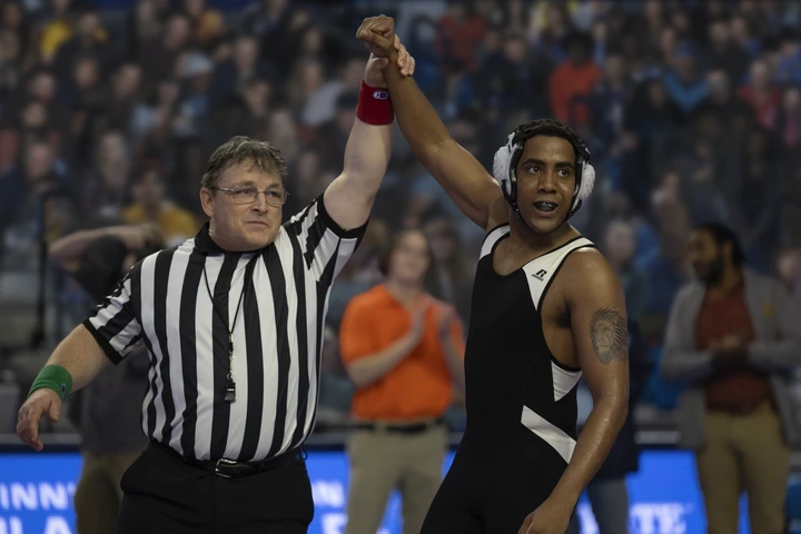 A referee holds up the arm of a victorious wrestler in a black and white singlet during a match, with a cheering crowd in the background