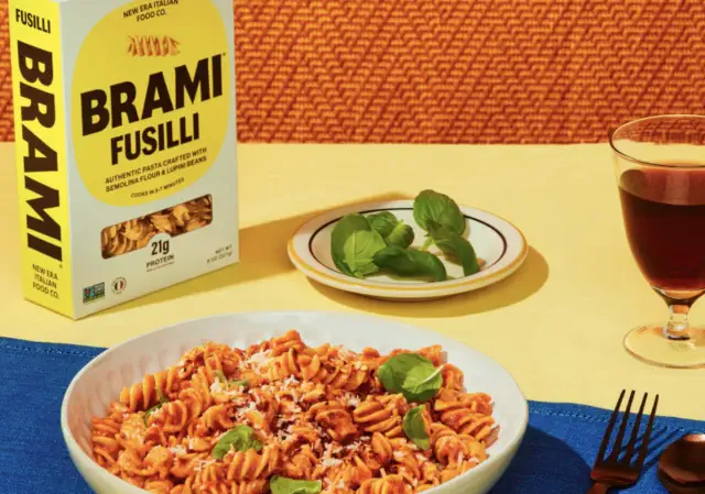 Brami fusilli pasta box next to a plate of cooked fusilli garnished with basil and cheese, with a drink and basil leaves on a table